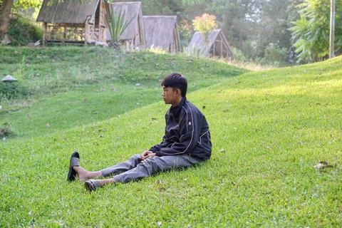 A boy sits alone on the green grass next to the remains of burnt garbage Stock Photos