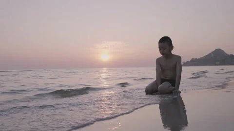 A boy sits on the beach with the sun reflect on the water behind Stock Footage 145977637