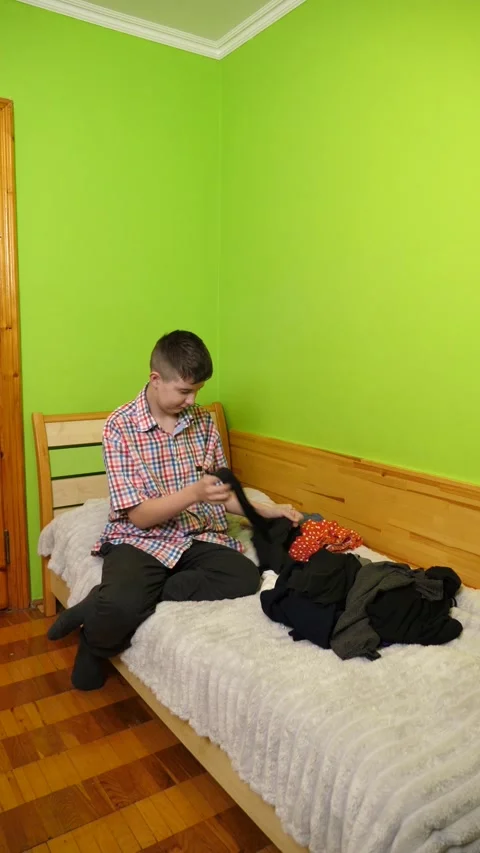A boy sits on the bed folding clothes, organizing his room and neatly arrangi Stock Footage 323367218