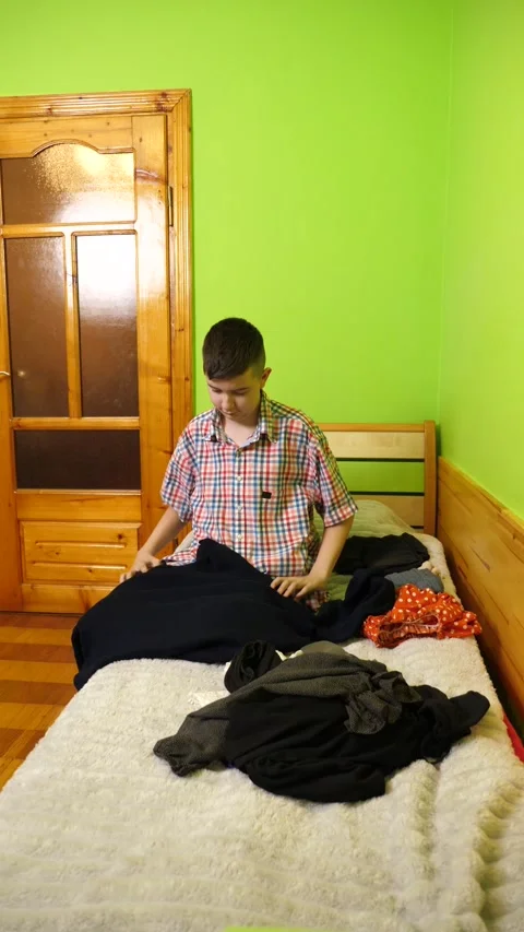 A boy sits on the bed folding clothes, organizing his room and neatly arrangi Stock Footage 323367292