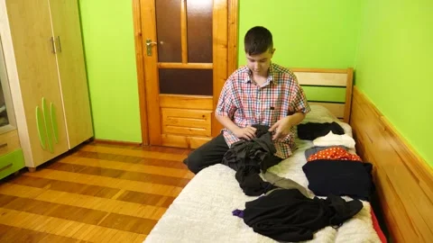 A boy sits on the bed folding clothes, organizing his room and neatly arrangi Stock Footage 323367293