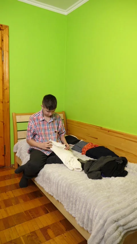 A boy sits on the bed folding clothes, organizing his room and neatly arrangi Stock Footage 323607293