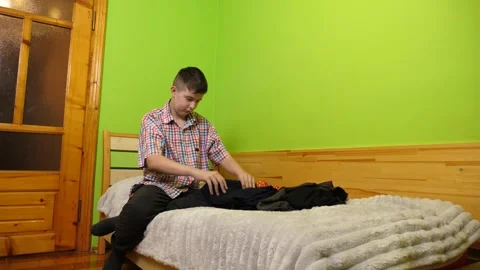 A boy sits on the bed folding clothes, organizing his room and neatly arrangi 動画素材 323607302