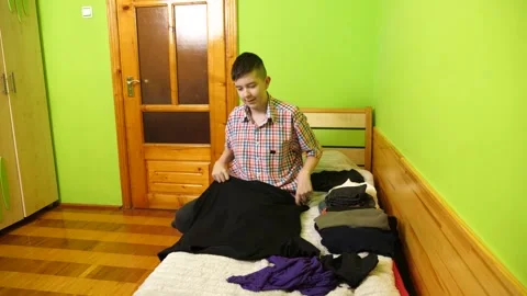 A boy sits on the bed folding clothes, organizing his room and neatly arrangi 動画素材 323607315