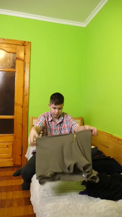 A boy sits on the bed folding clothes, organizing his room and neatly arrangi Stock Footage 323607327