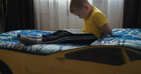 Boy sits on bed, tablet lies on his lap, hunched over looking at screen, games Stock Footage 123354332