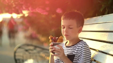Boy sits on a bench and eats ice cream wrapped in a waffle with chocolate and Stock Footage 261837472