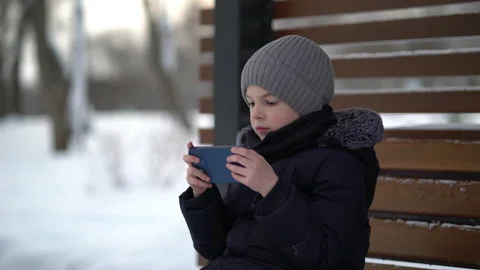 The boy sits on a bench in the park in winter and plays the phone. Stock Footage 234857301