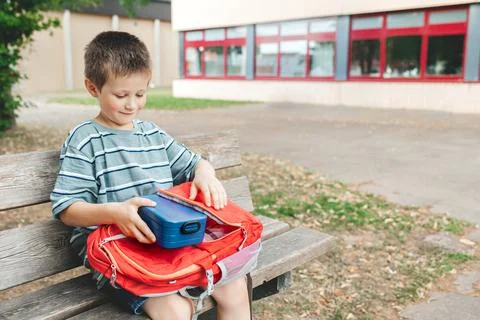 The boy sits on a bench in the school yard and takes out a lunch box from a Stock Photos