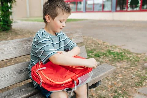 The boy sits on a bench in the school yard and takes out a lunch box from a Stock Photos