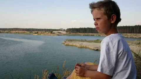 A boy sits on the edge of a cliff and looks into the distance at the lake Stock Footage 136876718