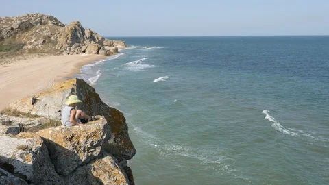Boy sits on the edge of the cliff. Waves wash the sandy shore. Ridge is broken Video stock 99111157