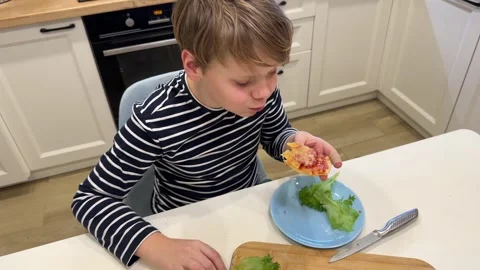 The boy sits at the kitchen table, eating pizza with enthusiasm Stock Footage 302927873