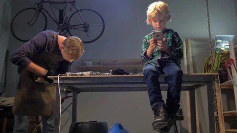 Boy sits with phone, master works with wood in a workshop Stock Footage 126721005