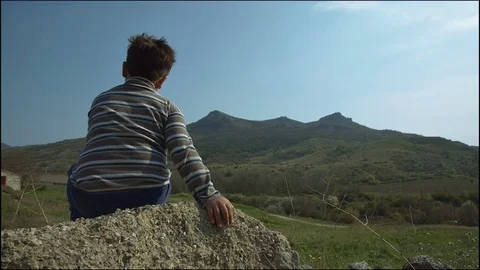 The boy sits on a rock in the background of a mountain ridge Stock Footage 75672573
