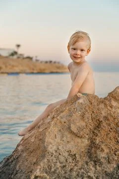 A boy sits on a rock, looking at the camera, with the sea in the background Stock Photos