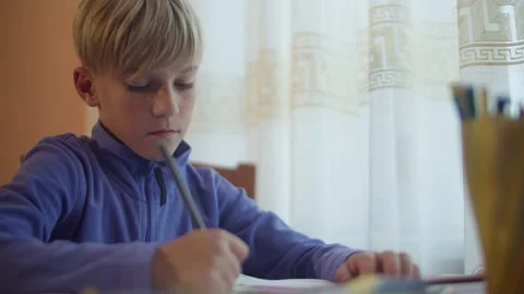 The boy sits at the table and draws with colored pencils.  Stock Footage 172057076
