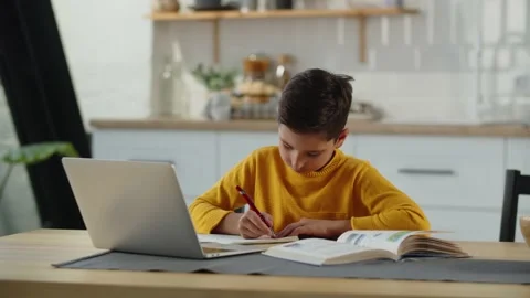 The boy sits at a table with a book and a laptop and writes something in a Stock Footage 240059659