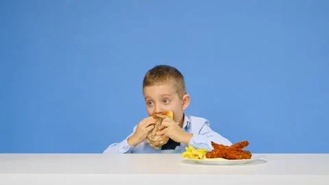 The boy sits at the table eats fast food and rejoices on a blue background. The Stock Footage 170996855