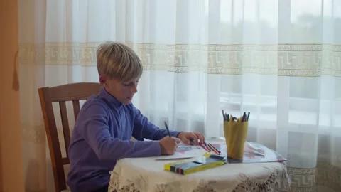 The boy sits at a table opposite the window and draws with pencils Stock Footage 172057077