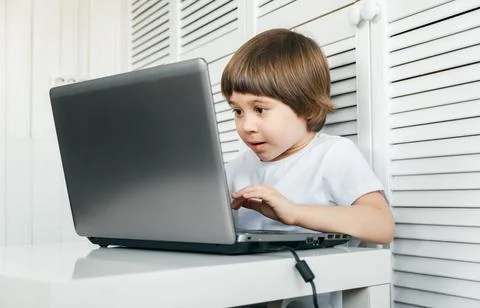 Boy sits at the table, uses laptop, typing on a keyboard, looks at the screen Stock Photos