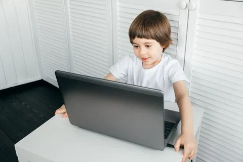 Boy sits at the table, uses laptop, typing on a keyboard, looks at the screen Stock Photos