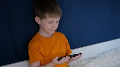 The boy is sitting on the bed with the phone in his hands, and makes funny faces Stock Footage 128204828