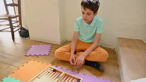 Boy sitting on floor playing backgammon in cozy home setting Stock Footage 290052704