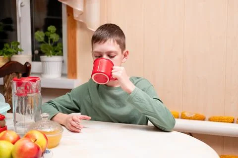 Boy sitting at the kitchen table and drinking a beverage from a bright red mug Stock Photos