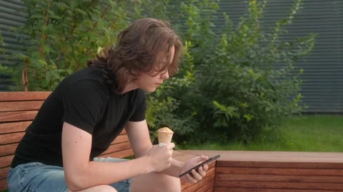 A boy is sitting on a park bench, eating ice cream and looking at his smartphone Video stock 299577671
