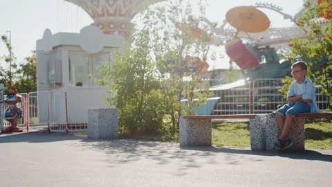 Boy sitting in park scenery. Young boy resting near carousel in sunlight. Young Stock Footage 333090438