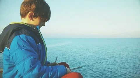 Boy is sitting on the pier looking at the sea and drawing Stock Footage 84234561