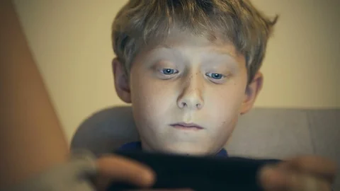 Boy sitting with portable digital device (phone or tablet) in hand, playing Stock-Footage 120265592