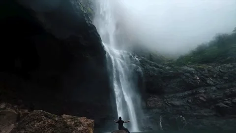 A Boy sitting on rock under waterfall and opening hands. Waterfall in forest Stock Footage 285929219