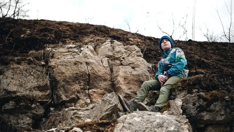 The boy is sitting on rocks on a steep slope near the river. Beautiful scenery Stock Footage 128016590