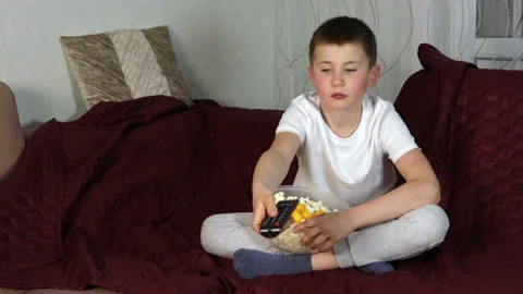 Boy sitting on sofa and eating popcorn and watching his favorite cartoons Stock Footage 154403546