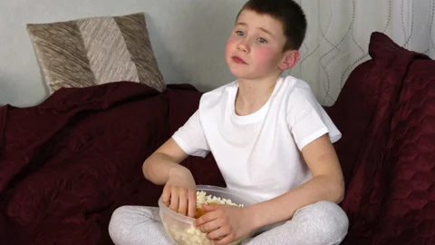 Boy sitting on sofa and eating popcorn and watching his favorite cartoons Stock Footage 154403760