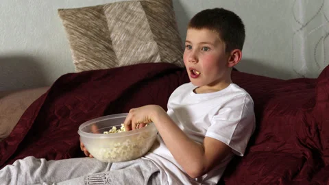 Boy sitting on sofa and eating popcorn and watching movie at home Stock Footage 154406726