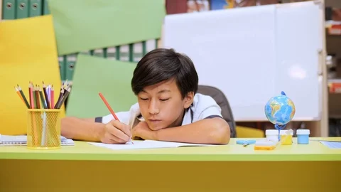 A boy sitting at a table and with his head on his hand writing something in a Video stock 93501421