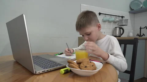 Boy is sitting at a table in front of a laptop, eating and drawing something Video stock 259681909