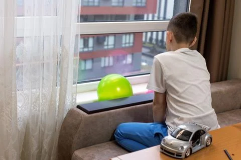 Boy sitting by window looking outside. A young boy in casual clothes sits Stock Photos
