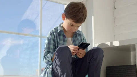 Boy sitting on windowsill, using mobile cell phone, watching video, playing game Stock Footage 171090700