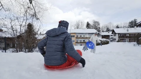 Boy sleds down snowy hill toward house past mother and brother Stock Footage 121754077
