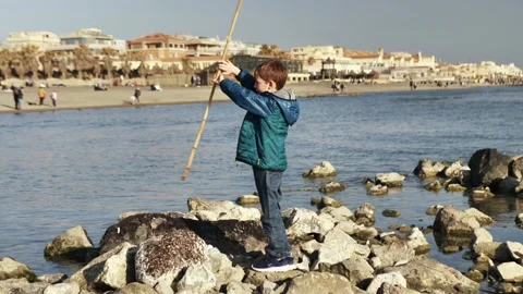 The boy spins a stick with his hands on the rocks by the sea. Video stock 150905976