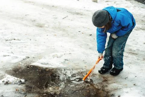 A boy standing in a blue jacket and hat breaking by stick ice in a frozen puddle Foto stock