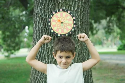 Boy standing in front of dart board on tree, arms up Stock Photos