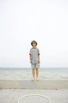 Boy standing on low wall at the beach, looking at camera Stock Photos