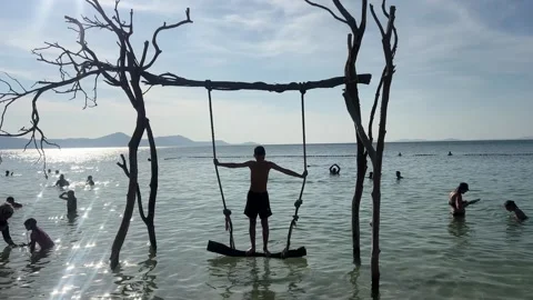 Boy Standing on a Rope Swing Over the Sea with People Swimming in the Background Stock Footage 336174670