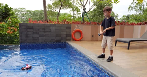 Boy stands by swimming pool, operating tiny tugboat with remote control, slow-mo Stock Footage 269497402