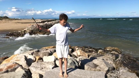 Boy staying on stone beach scaring by sudden sea wave splash Stock Footage 102329898